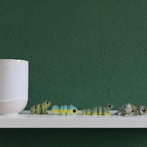 May include: A white ceramic mug sits next to four small articulated animal figurines including a green frog, a blue and yellow chameleon, a green lizard, and a grey turtle, followed by a small circular coin for scale on a white shelf.