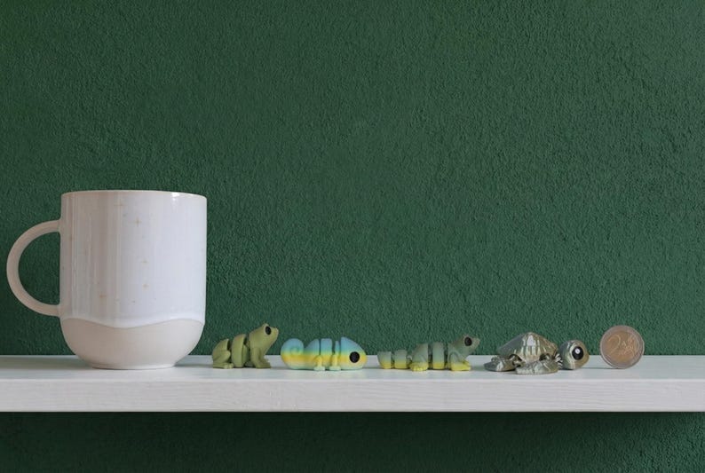May include: A white ceramic mug sits next to four small articulated animal figurines including a green frog, a blue and yellow chameleon, a green lizard, and a grey turtle, followed by a small circular coin for scale on a white shelf.