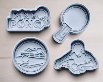 Op de afbeelding: Vier lichtblauwe koekjessnijders in verschillende vormen. Eén snijder spelt "PING PONG" met een pingpongbatje-pictogram. Andere snijders zijn een batje, een rond ontwerp met een net en bal en een speler.