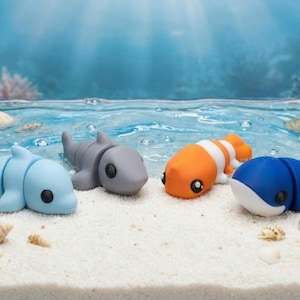 May include: Four articulated plastic sea creature figurines resting on a sandy surface. The set includes a light blue dolphin, a grey shark, an orange and white clownfish, and a dark blue whale, each featuring smooth surfaces and black eyes.