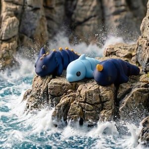 May include: Three small, smooth, articulated creature figurines resting on a rocky outcrop. One is dark blue with yellow spikes, one is light blue with a dorsal fin, and one is dark blue with a single yellow horn, all featuring large black eyes.
