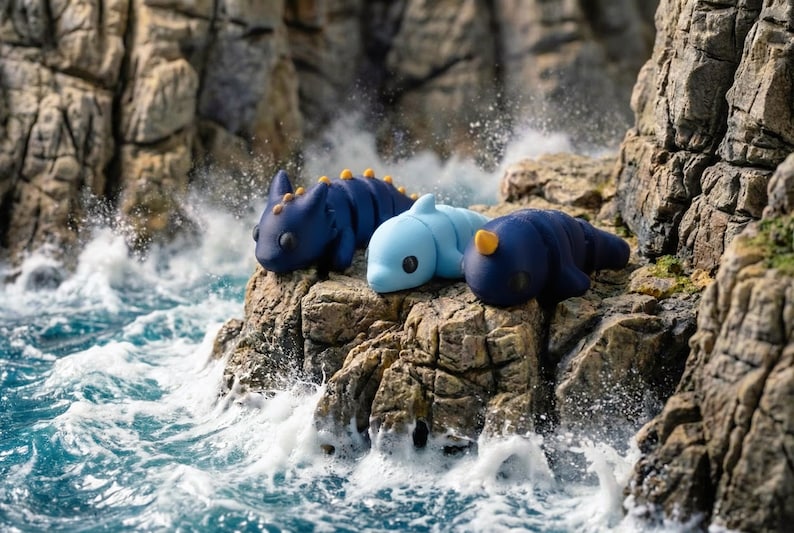 May include: Three small, smooth, articulated creature figurines resting on a rocky outcrop. One is dark blue with yellow spikes, one is light blue with a dorsal fin, and one is dark blue with a single yellow horn, all featuring large black eyes.