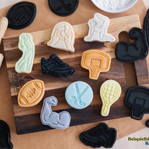 May include: A collection of black and white cookie cutters shaped like various sports equipment, including a football, basketball, tennis racket, hockey stick, and a weightlifting arm. The cookie cutters are arranged on a wooden surface with a rolling pin and several cookies decorated with fondant.