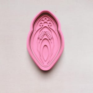 May include: A pink, layered cookie cutter in the shape of a stylized flower or botanical design. The cutter has a detailed floral pattern at the top and a central, elongated shape with layered details. The design is set against a neutral background.
