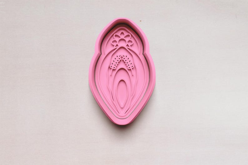 May include: A pink, layered cookie cutter in the shape of a stylized flower or botanical design. The cutter has a detailed floral pattern at the top and a central, elongated shape with layered details. The design is set against a neutral background.
