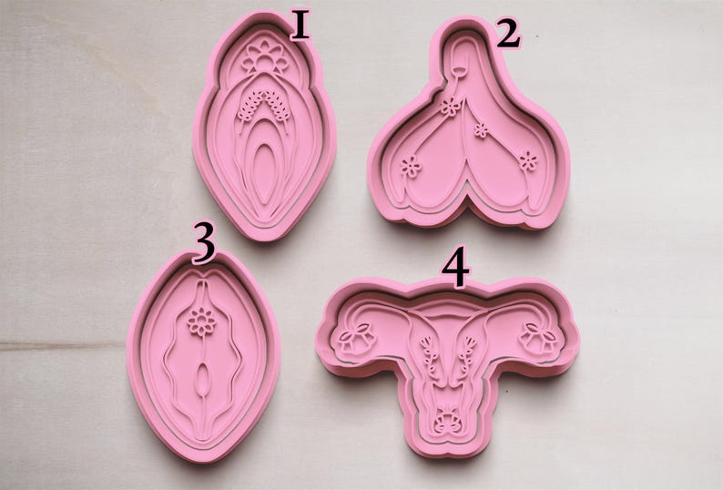 May include: Four pink cookie cutters in the shape of female reproductive organs. The cutters are made of a flexible material and are designed with detailed internal features. The shapes include a vulva, clitoris, and uterus.