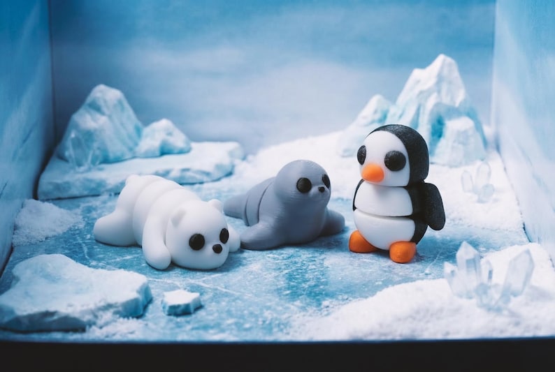 May include: A set of three small, smooth, rounded figurines featuring a white polar bear, a grey seal, and a black and white penguin with an orange beak and feet, arranged on a textured blue and white icy surface with miniature icebergs.
