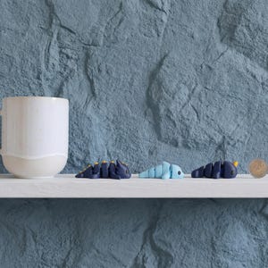 May include: A white ceramic mug sits next to three small, articulated plastic creature figurines in dark blue, light blue, and dark blue with yellow accents, positioned beside a silver coin for scale on a white shelf.