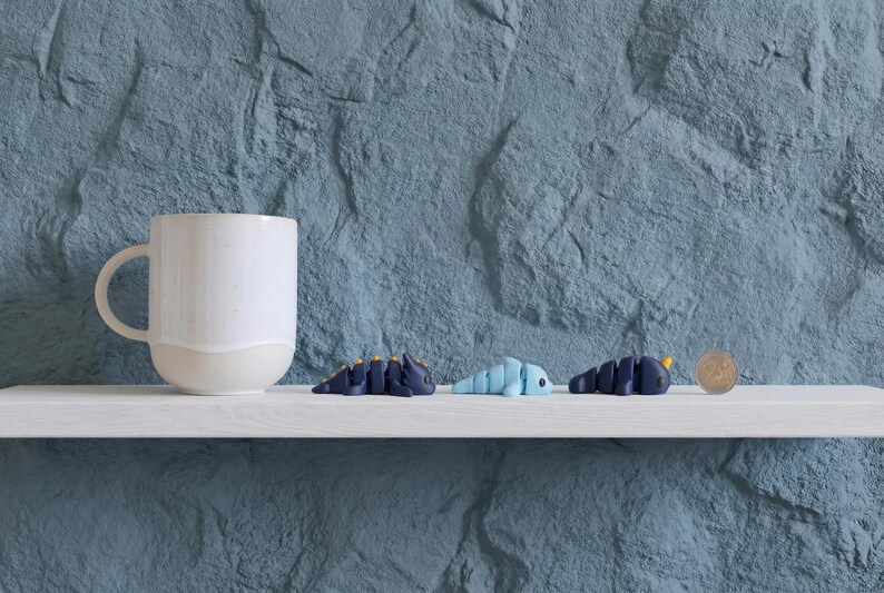 May include: A white ceramic mug sits next to three small, articulated plastic creature figurines in dark blue, light blue, and dark blue with yellow accents, positioned beside a silver coin for scale on a white shelf.