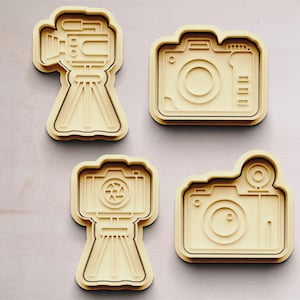 May include: Four wooden cookie cutters shaped like cameras. Two of the cutters are shaped like traditional cameras with a lens and a shutter button. The other two cutters are shaped like vintage cameras with a tripod.