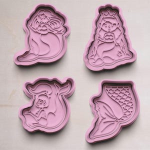 Puede incluir: Cuatro cortadores de galletas de plástico rosa con forma de sirenas. Los cortadores presentan una sirena con una corona, una sirena con el pelo largo, una cola de sirena y una sirena con una corona y una cola.