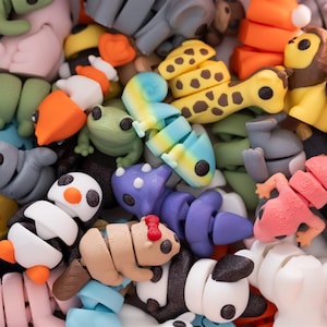 May include: A collection of articulated 3D printed animal figures in various colors and species, including a yellow giraffe with brown spots, a black and white penguin, a purple dinosaur, a pink lizard, a green frog, and a blue octopus.
