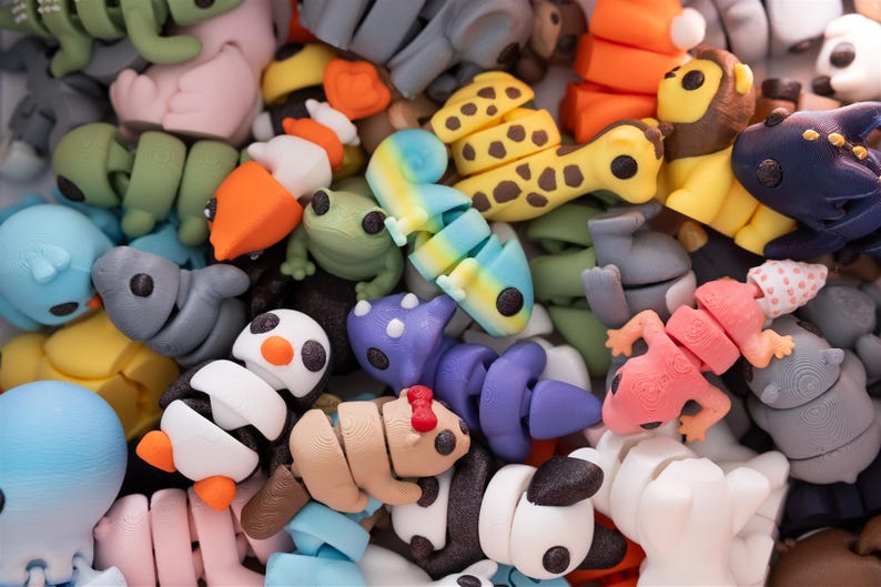May include: A collection of articulated 3D printed animal figures in various colors and species, including a yellow giraffe with brown spots, a black and white penguin, a purple dinosaur, a pink lizard, a green frog, and a blue octopus.