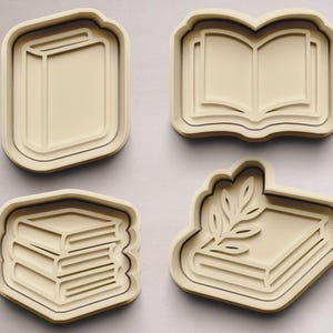 Könnte beinhalten: Vier beige Ausstecher in Buchform. Einer ist ein geschlossenes Buch, einer ein offenes Buch, einer ein Stapel Bücher und einer ein Buch mit Blättern. Diese sind wahrscheinlich zum Backen.