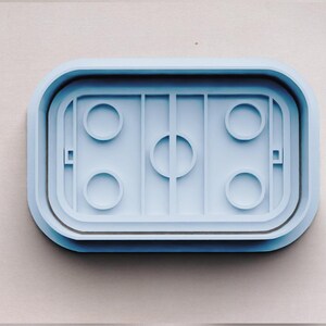 May include: A light blue plastic cookie cutter shaped like a hockey rink with four circles for cookies and a center circle.