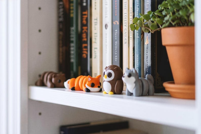 May include: A collection of four small animal figurines including a brown bear, an orange and white fox, a brown and white owl, and a gray husky dog, all arranged in a row on a white shelf in front of a series of books and a potted plant.