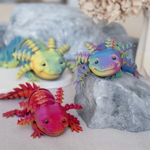 May include: Three colorful axolotl figurines with spiky details. The axolotls are posed on rocks and have a friendly, smiling expression. The axolotls are yellow, blue, and pink.