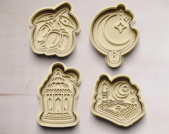 May include: Four cookie cutters in a light brown color. The cutters are shaped like a crescent moon with a star, a lantern, a date palm branch with dates, and a mosque with a crescent moon.