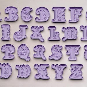 Puede incluir: Un juego de 26 cortadores de galletas de plástico morado con forma de las letras del alfabeto. Cada letra tiene un adorno decorativo.