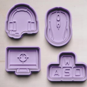 May include: Four purple silicone cookie cutters shaped like a headset, computer mouse, computer monitor with a target in the center, and a set of four keyboard keys labeled "WASD".