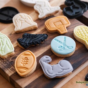 May include: A variety of sports-themed fondant cookies on a wooden cutting board. The cookies are decorated with images of a football, basketball, baseball, hockey, tennis, and a weightlifter. The cookies are all different colors and have a variety of designs.
