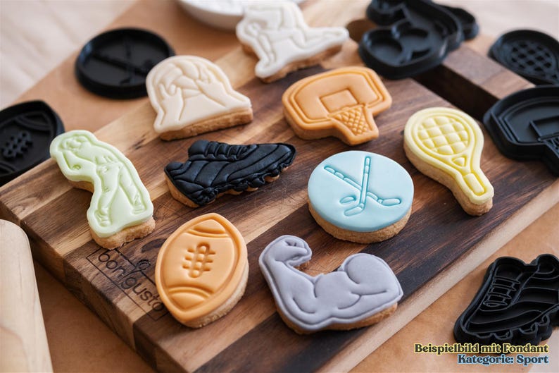 May include: A variety of sports-themed fondant cookies on a wooden cutting board. The cookies are decorated with images of a football, basketball, baseball, hockey, tennis, and a weightlifter. The cookies are all different colors and have a variety of designs.