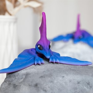 Op de afbeelding: Een paars en blauw dinosaurusbeeldje met grote vleugels. De dinosaurus ligt op een grijze rots.
