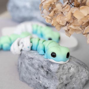 May include: A green and blue lizard figurine with a white belly and black eyes. The lizard is posed on a gray rock.