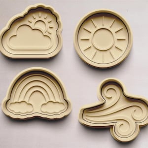 Puede incluir: Cuatro cortadores de galletas de color beige con forma de nube con sol, sol con rayos, arcoíris con nubes y remolino de viento.