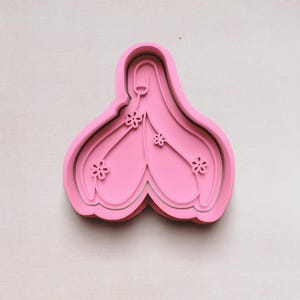 May include: A pink cookie cutter in the shape of a stylized flower with three smaller flower accents. The cutter has a layered design and is set against a white background.