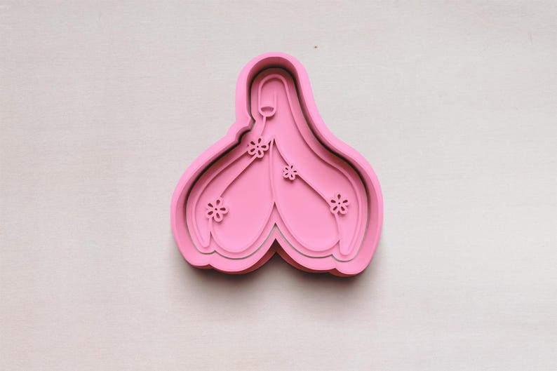 May include: A pink cookie cutter in the shape of a stylized flower with three smaller flower accents. The cutter has a layered design and is set against a white background.
