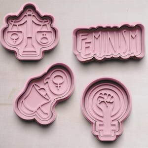 May include: Four pink plastic cookie cutters, each with a different feminist symbol. The cutters are shaped like a scale, a fist in a circle, a bell, and the word "FEMINISM".