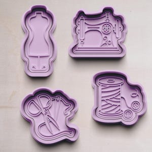 Könnte beinhalten: Vier lavendelfarbene Ausstecher in Form einer Schneiderpuppe, einer Nähmaschine, einer Schere mit Faden und einer Garnrolle mit Knöpfen. Ideal für thematische Kekse für Näh- oder Modebegeisterte.