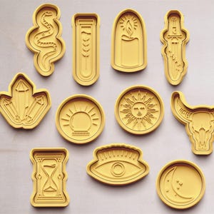 Könnte beinhalten: Set mit 10 gelben Ausstechern mit verschiedenen Designs, darunter eine Schlange, ein Kristall, eine Sonne, ein Auge, eine Mondsichel, ein Stierkopf, eine Sanduhr, ein Reagenzglas, eine Kerze und ein Dolch.