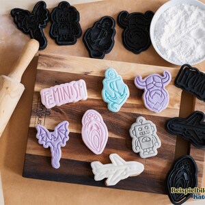 May include: A collection of decorated cookies and cookie cutters on a wooden cutting board. The cookies feature various shapes, including a robot, airplane, anchor, and the word "Feminism". A rolling pin and a bowl of white powder are also present.