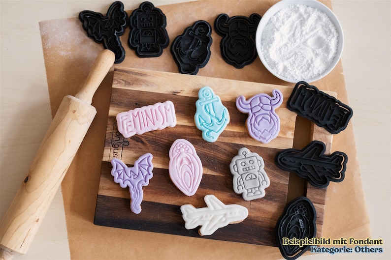 May include: A collection of decorated cookies and cookie cutters on a wooden cutting board. The cookies feature various shapes, including a robot, airplane, anchor, and the word "Feminism". A rolling pin and a bowl of white powder are also present.