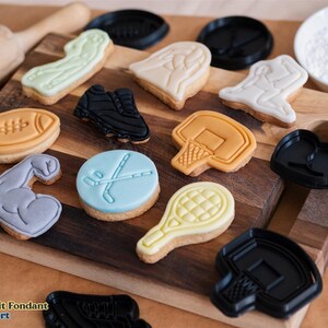 May include: A variety of cookie cutters in different sports themes, including football, basketball, hockey, tennis, and weightlifting. The cutters are made of black plastic and are arranged on a wooden surface.  The image also shows cookies decorated with fondant in the shapes of the cutters.