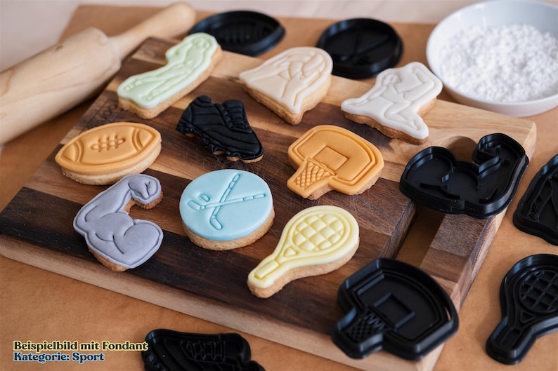 May include: A variety of cookie cutters in different sports themes, including football, basketball, hockey, tennis, and weightlifting. The cutters are made of black plastic and are arranged on a wooden surface.  The image also shows cookies decorated with fondant in the shapes of the cutters.