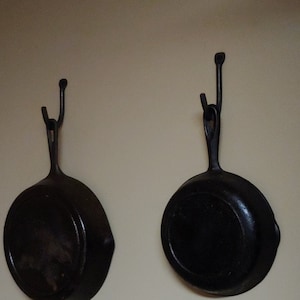 May include: Two black cast iron skillets hanging on hooks against a light beige wall.