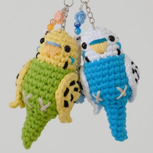 May include: Two crocheted parakeet keychains, one green and yellow, the other blue and white. Both have black eyes and a white stitched X on their bellies.