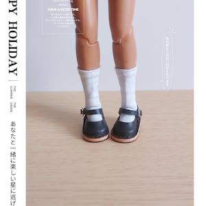 Pode incluir: Sapatos pretos de boneca com tira e fivela, combinados com meias brancas. As pernas da boneca são visíveis, em pé sobre uma superfície de madeira. O texto japonês está incluído.