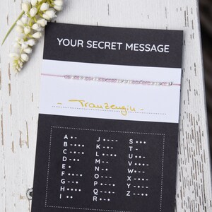 Op de afbeelding: Een zwarte kaart met de tekst "YOUR SECRET MESSAGE" en een roze en witte kralen armband. De kaart bevat ook een codetabel met letters van het alfabet en hun bijbehorende morsecode.