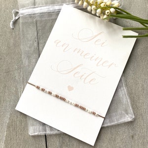 May include: A white card with the text "Sei an meiner Seite" in pink script. A small pink heart is below the text. A brown and white beaded bracelet is on the card.