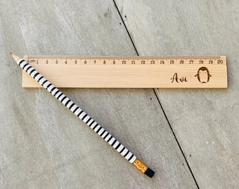 May include: A wooden ruler with a penguin design and the name "Avi" engraved on it. The ruler is 20 cm long and has black and white stripes on the back.