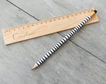 May include: A wooden ruler with a black and white striped pencil. The ruler is marked with centimetres and numbers 0 to 20. The ruler is personalised with the name "Emilia".