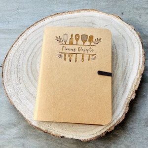 May include: A brown recipe journal with a wooden cover. The cover has a design of kitchen utensils and the text "Fiona's Rezepte".
