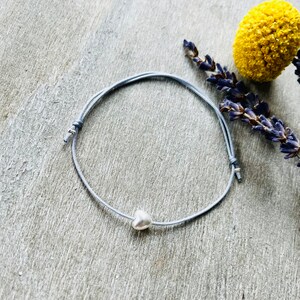 Pode incluir: Um delicado bracelete de corda cinza com um pequeno berloque de coração prateado.
