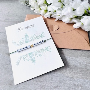 Könnte beinhalten: Eine Grußkarte mit der Aufschrift "Für meine" und einem Armband mit den Buchstaben "BRAUT MAMA" aus weißen Perlen und einem goldenen Herz. Die Karte hat ein florales Design und liegt neben einem braunen Umschlag und weißen Blumen.