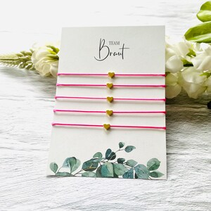 Könnte beinhalten: Ein Set mit sechs rosafarbenen Freundschaftsarmbändern mit goldenen Herzanhängern. Die Karte im Hintergrund lautet "Team Braut".