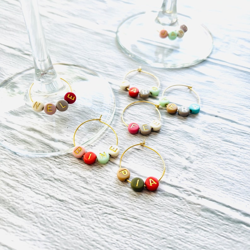 May include: Six gold-colored wine glass charms with colorful beads spelling out the names "Nele", "Bine", "Fe", "Pia", "Caro", and "Zer".